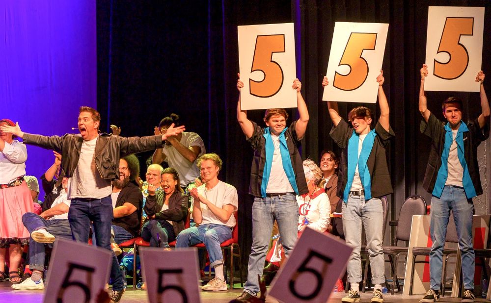 Actors on stage with 3 men holding up signs showing the number 5