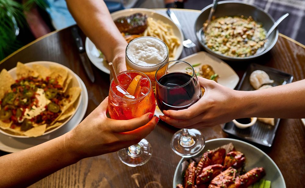 Three people toasting with beer, red wine and a spritz over shared plates at Hotel Coronation