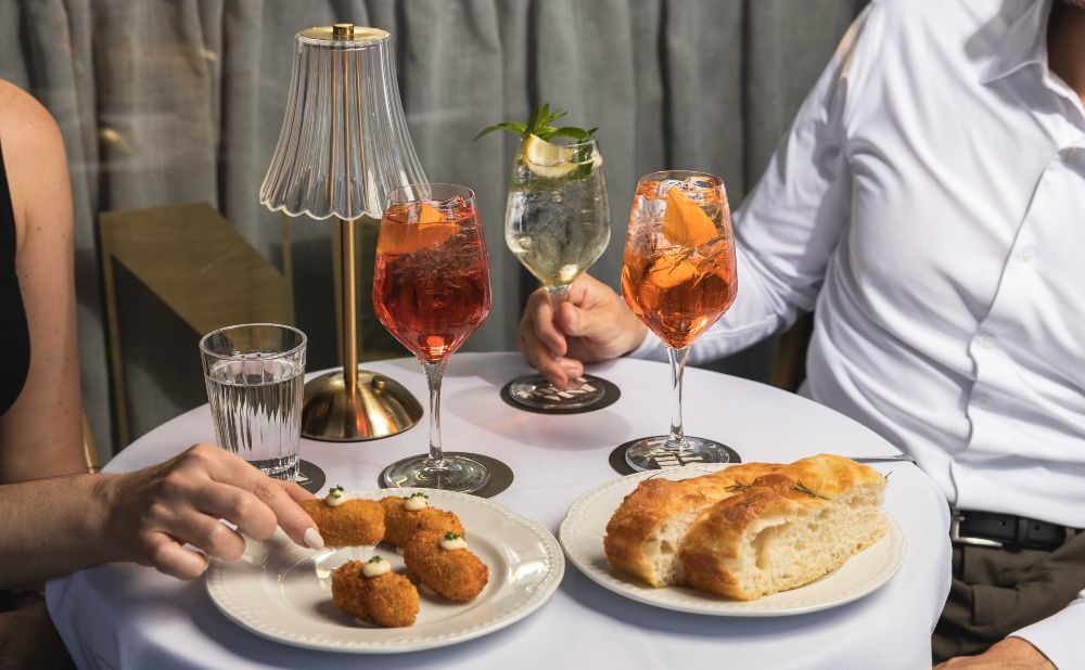 Two people toastingwith Aperol spritz and a white wine spritz outdoors, with croquettes on the table