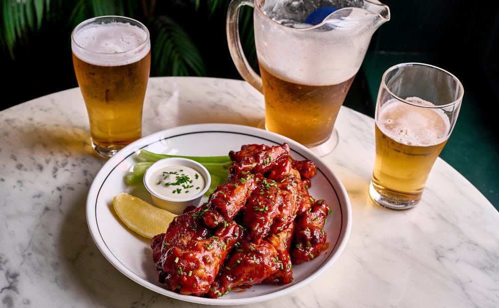 Plate of saucy chicken wings with celery and ranch beside a jug and two glasses of beer at Keg & Brew