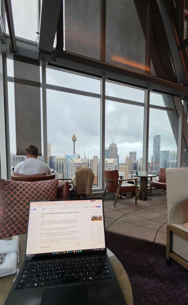 Working at a table in Club Millésime lounge with Sydney skyline views including Sydney Tower and CBD buildings visible through floor-to-ceiling windows
