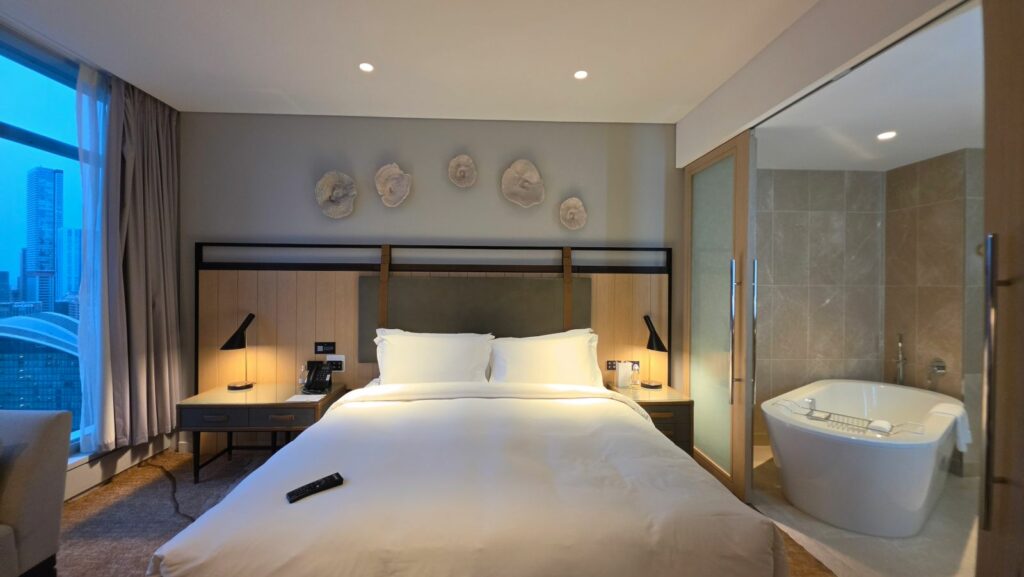 Sofitel Sydney Darling Harbour hotel room showing king bed with white linens, decorative fossil art on wall, freestanding bathtub visible through open bathroom area, and city views through window